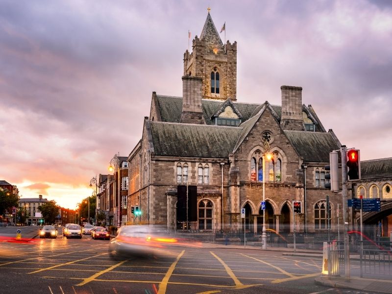 A historic stone cathedral with a tall central tower stands at a busy city intersection at dusk. Blurred headlights from passing cars streak across the foreground, and traffic lights and streetlamps illuminate the scene under a cloudy sky.