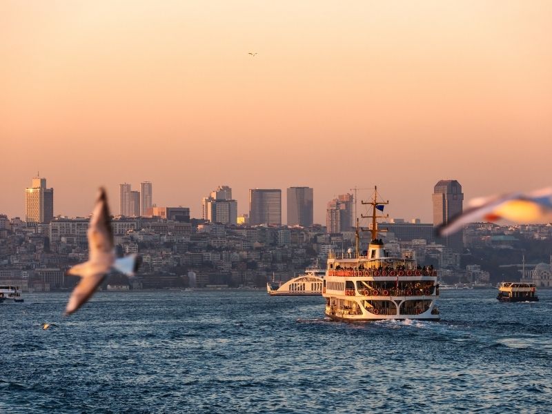 Ferry glides through a bustling waterway at sunset, city skyline in the background.