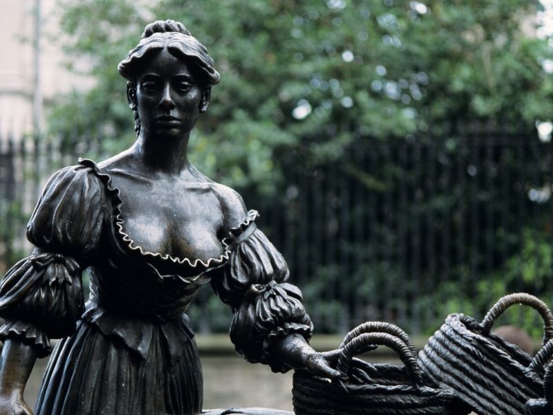 Bronze statue of a woman with braided hair in period dress, holding baskets. Set against a blurred backdrop of greenery and an iron fence.