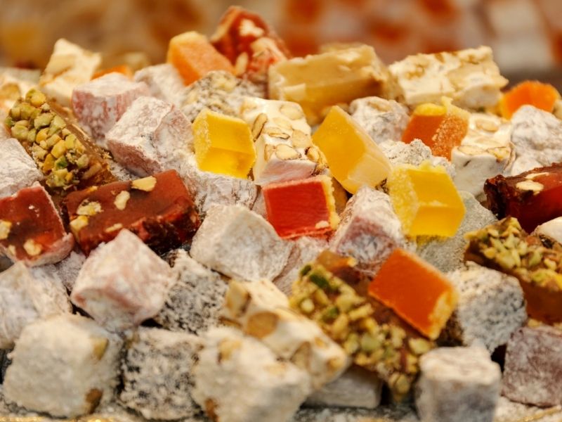 A vibrant assortment of Turkish delight cubes, in various colours like orange, yellow, and red, dusted with powdered sugar and nuts.