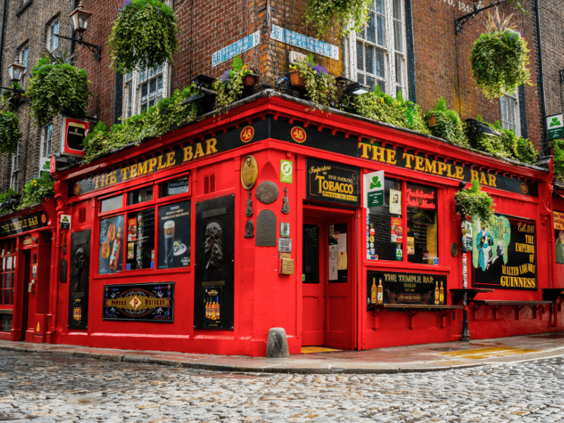 Bright red pub with hanging green plants, named "The Temple Bar." Alley setting, cobblestone streets. Colorful, lively atmosphere.