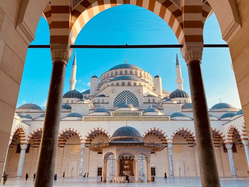 A majestic mosque with ornate domes and minarets is framed by archways. The bright blue sky enhances the grandeur, evoking a serene, awe-inspiring atmosphere.
