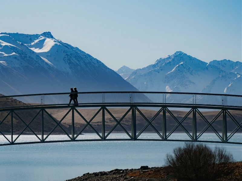 couple walking over bridge with snow capped mountains in background