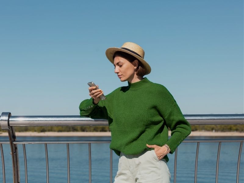 stylish woman wearing green casual sweater hat outdoor bridge with river view holding mobile phone